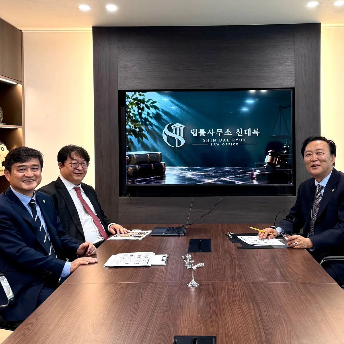 법률사무소 신대륙 대표변호사 단체 사진. 사무소 로고가 띄워진 회의실 화면 앞, 회의 테이블에 세 사람이 둘러앉아 있습니다.